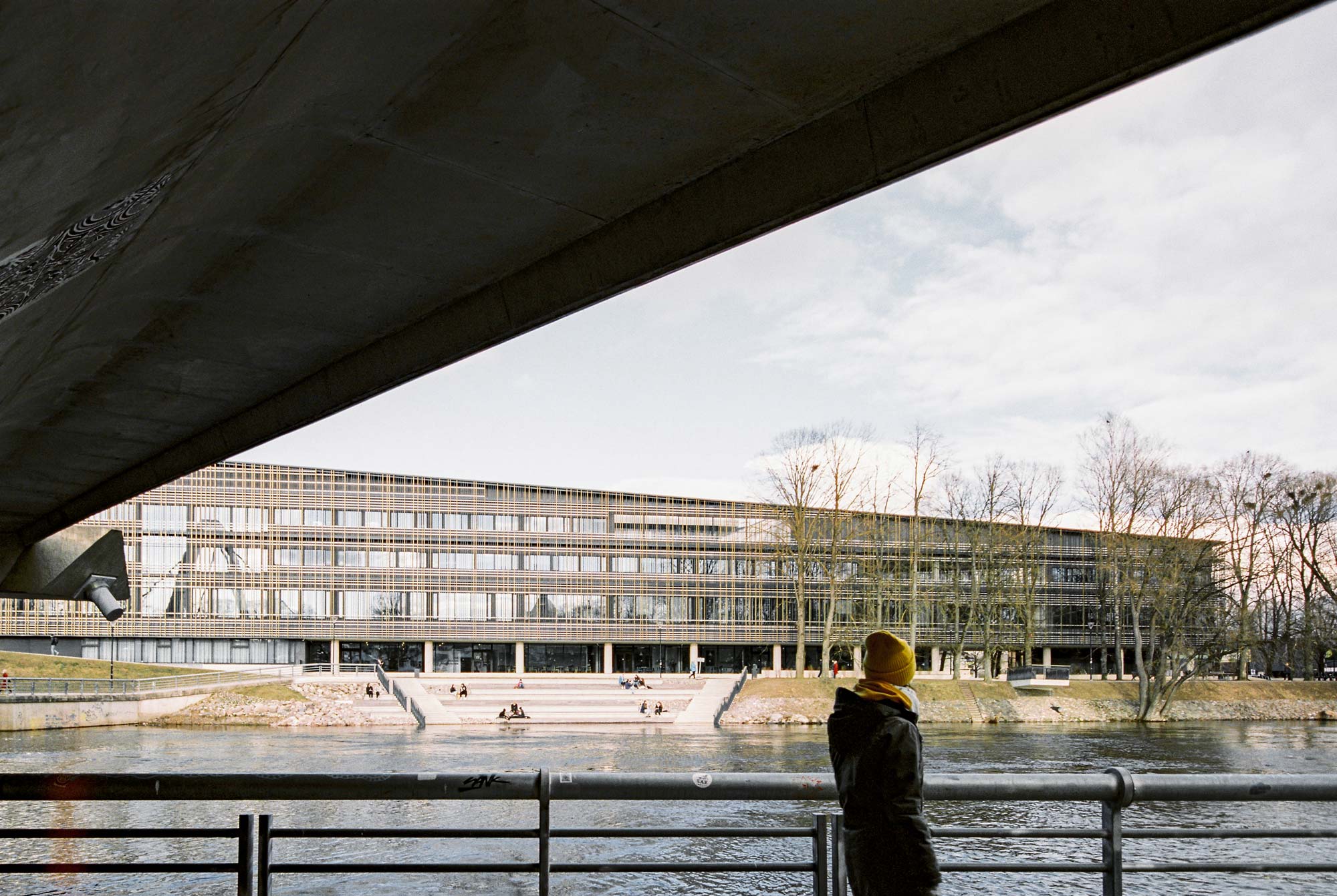 Man walking under a bridge with river flowing and large building accross with people seating in front